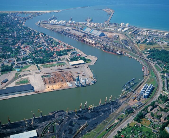 Aerial view of Ventspils industrial harbour, showcasing cargo ships docked at the extensive piers, with multiple cranes and silos along the quayside. The image captures the port's infrastructure, including construction areas with raw materials. Adjacent to the port is the residential area of Ventspils, blending into the urban setting. The port is protected from the open sea by a significant breakwater, facilitating safe and efficient maritime operations.