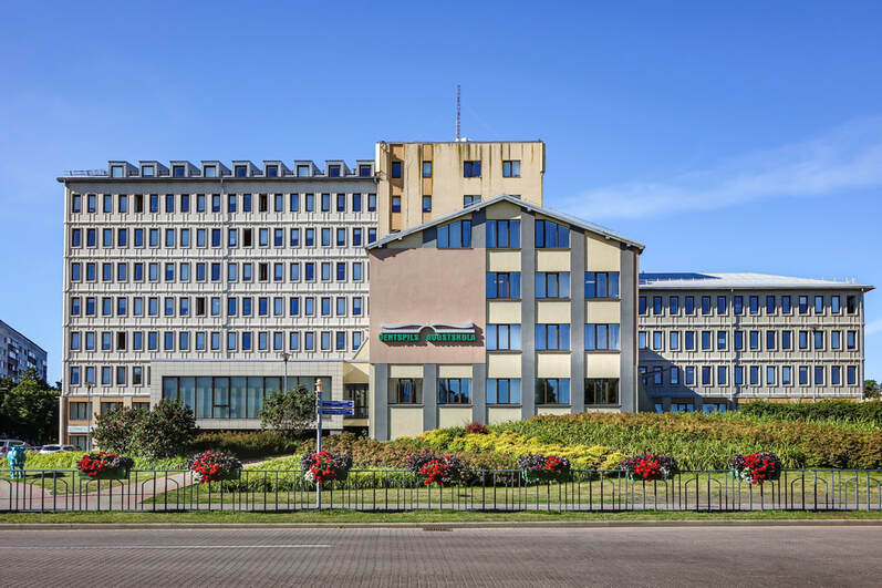 Facade of the Ventspils Augstskola (Ventspils University College), with a modern multi-story building adorned with flowers and landscaped greenery in the foreground.