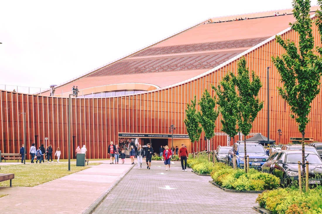 Exterior view of the Ventspils Science Centre, Vizium, featuring modern architecture with a distinctive curved, rust-coloured facade. People are seen entering and exiting the building, with vibrant green trees lining the approach. A parking area is partially visible to the right, with cars parked alongside the landscaped pathway.